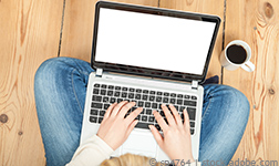 Blonde woman typing on laptop sitting on wooden floor with cup of coffee next to her