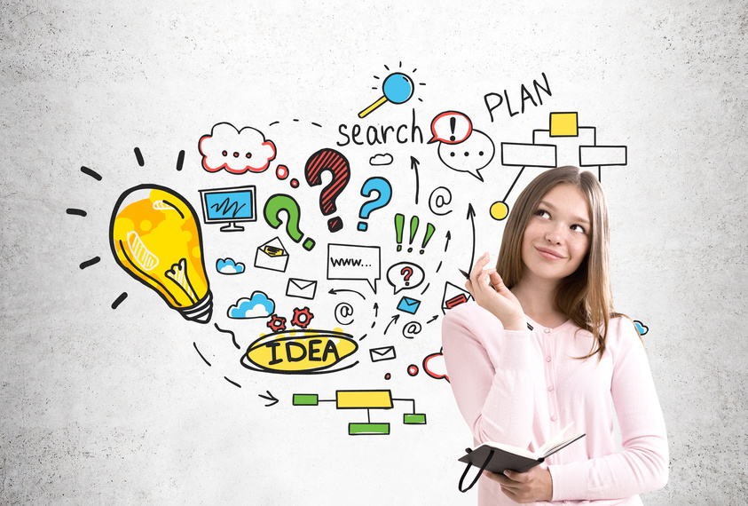 Portrait of a young woman wearing a pink cardigan and holding a notebook while standing near a concrete wall with a colorful business plan sketch