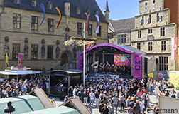 A large crowd gathers in front of a stage where a choir is performing. The stage is decorated with colorful posters and lights. Historical buildings can be seen in the background. The weather is bright and the atmosphere is festive.